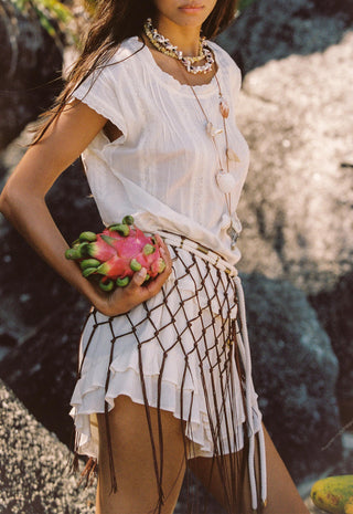 A woman sits on a sandy beach wearing a white short-sleeve top, a white skirt with a brown net pattern, and layered necklaces. Coconuts are visible in the foreground, and tropical foliage and rocks are in the background.
