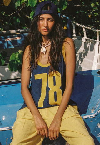 A woman wearing a navy blue athletic tank top with yellow trim and the number 78, paired with yellow pants, a navy cap, and a chunky silver necklace, sitting on a weathered blue vehicle outdoors.