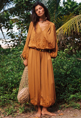 A woman stands outdoors in a lush green tropical setting wearing a matching rust-orange bohemian two-piece set consisting of a long-sleeve embroidered top and wide-leg pants. She is barefoot and holds a woven straw bag.