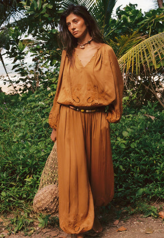 A woman stands outdoors in a lush green setting wearing a matching rust-brown bohemian long-sleeve top and wide-leg skirt set with a black belt. She is holding a woven straw bag.