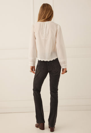 a-woman-viewed-from-behind-wearing-a-cream-colored-long-sleeve-blouse-with-lace-trim-and-dark-charcoal-gray-jeans-standing-against-a-white-paneled-wall