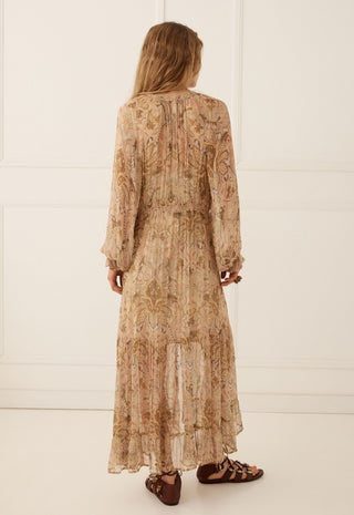A woman viewed from behind wearing a long-sleeved, midi-length beige maxi dress with an intricate paisley pattern in warm brown and tan tones. She is wearing brown strappy flat sandals against a clean, white paneled wall background.