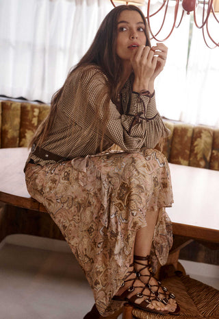 a-woman-sits-on-a-wooden-bench-in-a-warm-sunlit-room-wearing-a-sheer-patterned-brown-and-beige-blouse-with-a-matching-long-skirt-lace-up-strappy-sandals-and-bracelets
