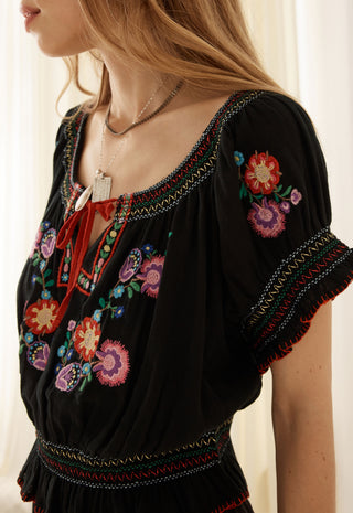 a-close-up-of-a-woman-wearing-a-black-bohemian-style-blouse-with-colorful-floral-embroidery-and-a-red-tie-neck-detail-paired-with-layered-silver-necklaces