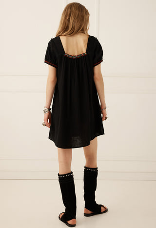 A woman viewed from behind wearing a loose black short-sleeve dress with a decorative embroidered neckline, black studded leg warmers, and black sandals, standing against a white paneled wall.