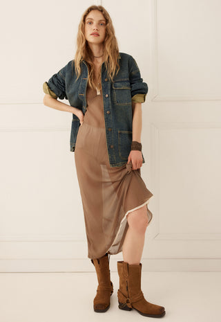 a-woman-models-a-brown-midi-slip-dress-with-a-sheer-overlay-styled-with-a-blue-denim-shirt-jacket-brown-leather-boots-and-a-brown-choker-against-a-white-paneled-wall