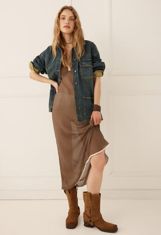 a-woman-models-a-bohemian-style-outfit-consisting-of-a-brown-midi-slip-dress-a-blue-denim-jacket-and-brown-suede-boots-set-against-a-clean-white-paneled-wall