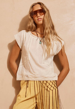 A woman wearing a white sleeveless top with vertical textured stripes and yellow fringe pants, posing against a textured beige wall.