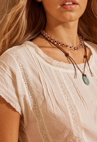 A close-up of a woman wearing a cream-colored lace-trimmed blouse and layered necklaces, including a brown cord necklace with a green pendant.