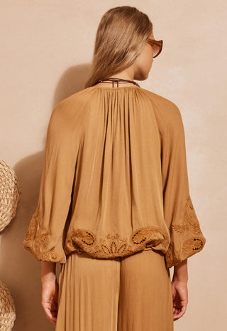 A woman shown from the back wearing a camel brown pleated blouse with embroidered lace sleeves and matching pleated trousers, set against a warm beige textured wall.