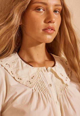 A close-up shot of a woman wearing a cream-colored blouse with a large eyelet lace Peter Pan collar and smocked detailing on the chest.