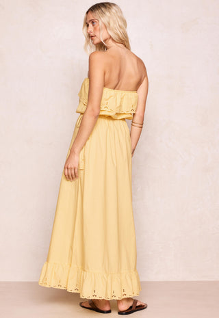 A woman modeling a pale yellow strapless maxi dress with ruffle details, shown from the back against a textured beige wall.