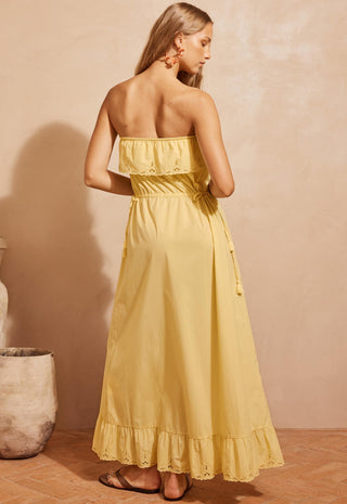 A woman modeling a strapless yellow maxi dress with ruffle details, standing against a textured beige wall in a warm, rustic setting.