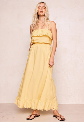 A woman modeling a pale yellow strapless maxi dress with ruffle details, paired with brown sandals, standing against a textured beige wall.