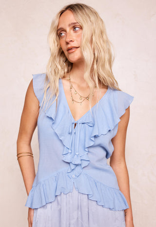 A blonde woman wearing a light blue sleeveless ruffled blouse with a tie-front detail and a matching light blue skirt, posed against a neutral beige background.