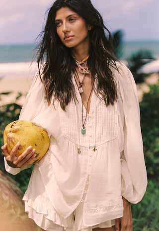 a-woman-on-a-beach-wearing-a-loose-white-bohemian-style-blouse-and-holding-a-yellow-coconut-with-layered-necklaces-and-a-tropical-ocean-background