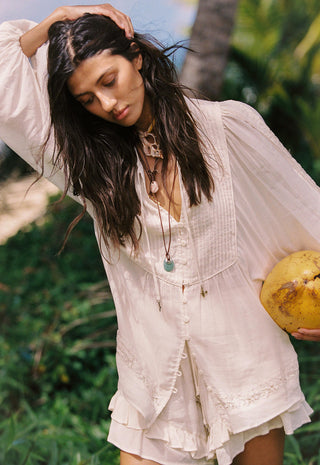 a-woman-outdoors-wearing-a-loose-light-cream-boho-style-blouse-with-long-sleeves-and-a-ruffled-hem-holding-a-yellow-coconut-she-is-surrounded-by-green-foliage-and-a-palm-tree-in-a-tropical-setting