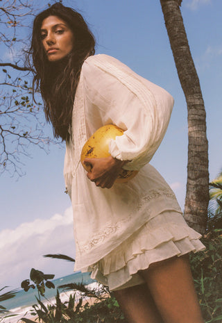 A woman wearing a loose white long-sleeve mini dress with lace trim stands outdoors near a palm tree