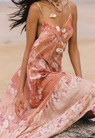 a-woman-seated-on-a-beach-wearing-a-flowing-floral-maxi-dress-in-soft-peach-and-pink-tones-with-white-flower-motifs-the-dress-has-thin-straps-and-a-backless-open-design-with-shell-like-embellishments