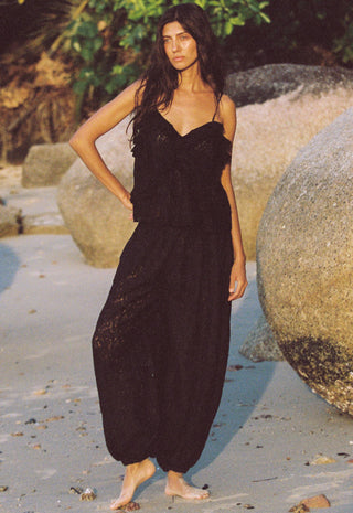 A woman stands barefoot on a sandy beach with large rocks and greenery in the background
