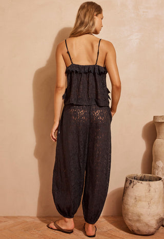 A woman stands with her back to the camera wearing a black lace camisole top with thin straps and matching black lace harem pants, photographed in a warm beige studio setting with decorative vases.