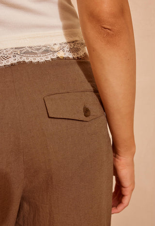 Close-up of a person wearing brown wide-leg trousers with a flap pocket and button, paired with a cream lace-trim top against a warm beige background.