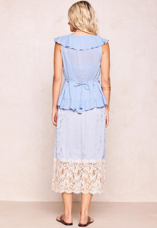 A woman shown from the back wearing a light blue ruffled midi dress with a lace hem, standing against a neutral beige wall.