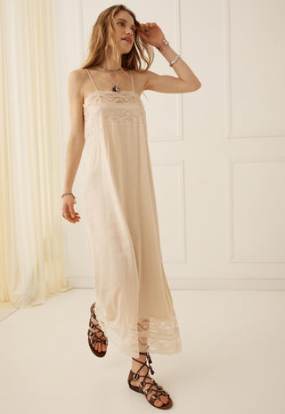 A woman models a long cream-colored slip dress with delicate lace trim at the bodice and hem, paired with brown strappy flat sandals in a bright, minimalist room with white paneled walls and a sheer cream curtain.
