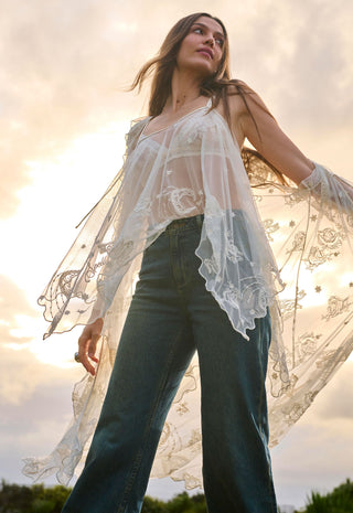 a-woman-wearing-a-sheer-white-floral-lace-long-sleeve-top-over-a-white-camisole-and-dark-blue-wide-leg-jeans-photographed-outdoors-against-a-bright-sky-with-the-sun-shining