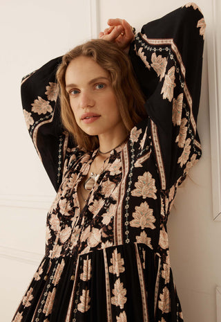 A woman wearing a black bohemian-style maxi dress with beige floral embroidery and wide bell sleeves, posing against a white paneled wall.