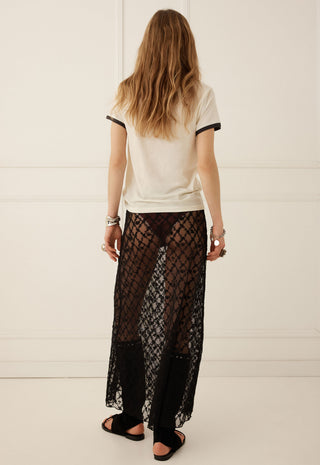 A woman viewed from behind wearing a cream ringer t-shirt with black trim and a sheer black lace maxi skirt, standing against a white paneled wall.